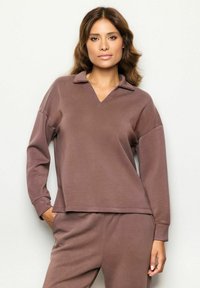 Mujer con cabello castaño ondulado vestida con un sudadera de cuello y mangas largas en color malva y pantalones a juego, de pie con una mano en el bolsillo.