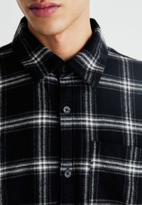 Camicia in flanella a quadri bianca e nera con colletto, chiusura a bottoni e taschino sul petto. Il tessuto appare morbido e con una texture.