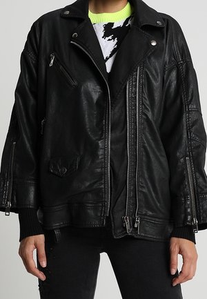 Veste en cuir noire oversize avec plusieurs zips et boutons-pression, portée sur un pull imprimé noir et blanc avec un col vert néon.