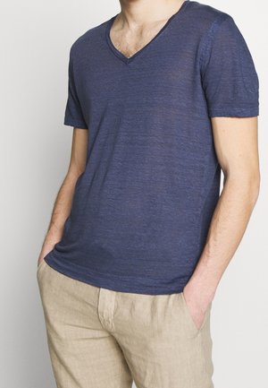 Homme portant un t-shirt bleu marine semi-transparent à col en V et un pantalon beige, les mains dans les poches, torse visible du cou jusqu'aux hanches.