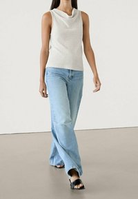 Woman walking in sleeveless white top, light blue wide-leg jeans, and black open-toe sandals against plain background.