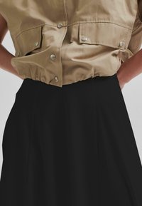Khaki korte mouwen top met twee drukknop-flapzakken, geplooide zoom en zilveren hardware, gecombineerd met een vloerlange zwarte rok.