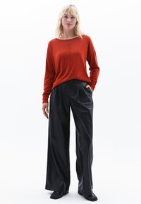 Rotes langärmliges Strickoberteil mit weitem Ausschnitt, kombiniert mit schwarzen hochgeschnittenen weiten Hosen aus glänzendem Material.
