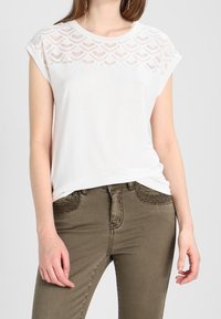 Maglietta bianca con un inserto in pizzo testurizzato, maniche corte e una vestibilità rilassata, abbinata a jeans verde oliva con dettagli decorativi sulle tasche.