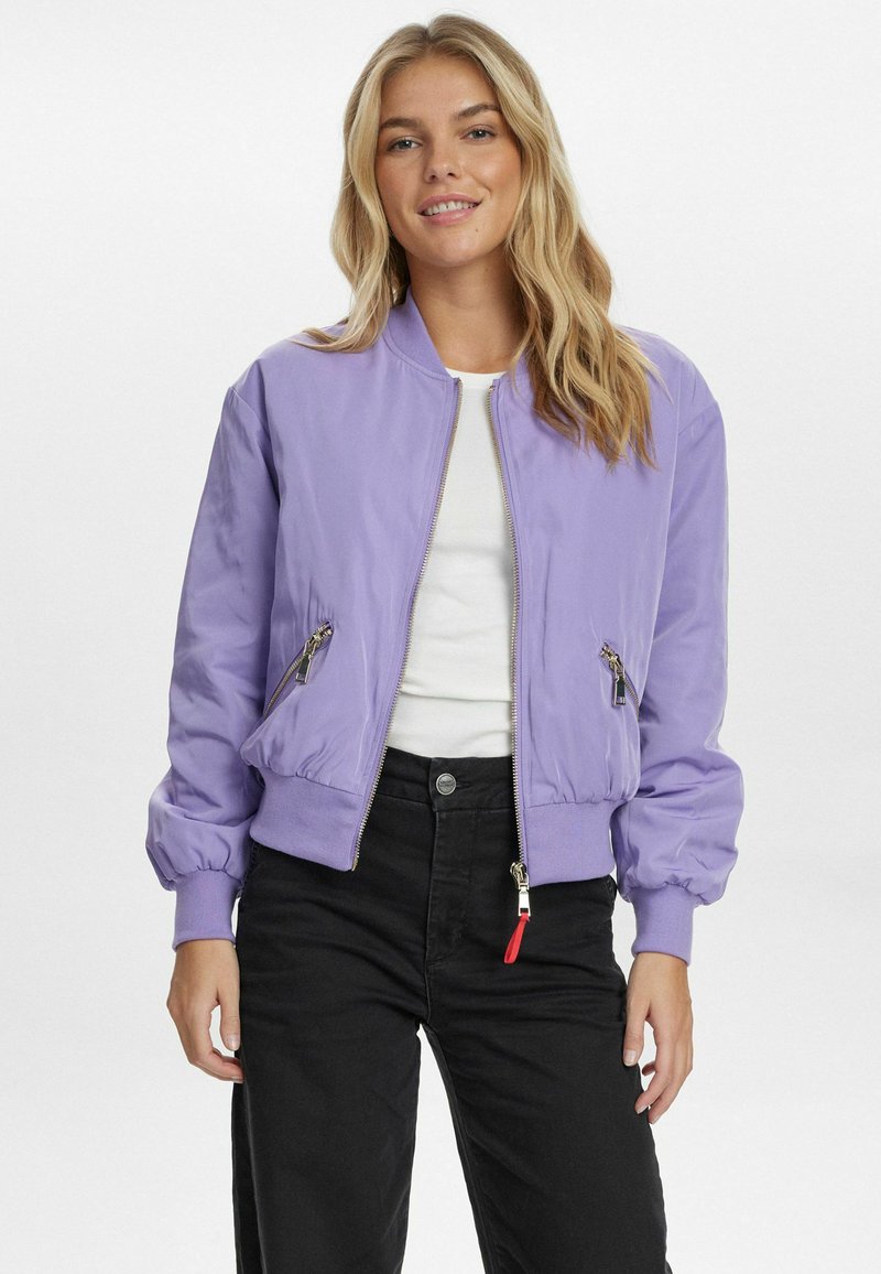 Lilac krāsu bomber jaka ar rāvējslēdzēju priekšpusē, ribotām piedurknēm un sānu kabatām; izgatavota no spīdīga auduma un ar kontrastējošiem rāvējslēdzējiem. Saskaņota ar melniem biksēm.