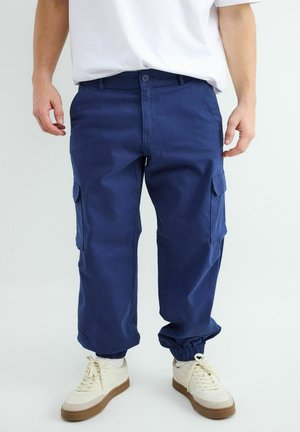 Man draagt marineblauwe cargobroek met elastische manchetten en witte sneakers, staande tegen een effen achtergrond.