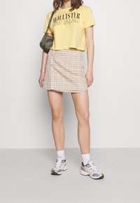 Žluté zkrácené tričko s černým potiskem, béžová mini sukně s kostkovaným vzorem a předními knoflíky, bílé kotníkové ponožky a šedé tenisky s akcenty.