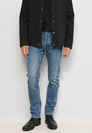 Persona que lleva vaqueros ajustados azul claro, cinturón negro, camisa negra abotonada y botas negras con cordones, de pie frente a un fondo liso.