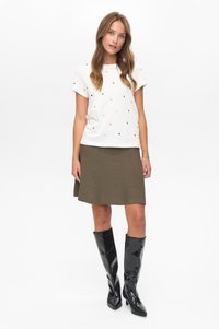 Junge Frau steht, trägt ein weißes kurzärmliges Hemd mit Punkten, einen olivgrünen Rock und schwarze kniehohe Stiefel vor einem schlichten weißen Hintergrund.