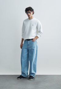 Λευκό φούτερ με μικρό μαύρο λογότυπο, συνδυασμένο με ανοιχτού μπλε χρώματος τζιν παντελόνι με φαρδιά μπατζάκια και μαύρα αθλητικά παπούτσια. Το μοντέλο φορά γυαλιά. Απλό φόντο.