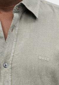 Κοντινό πλάνο ενός ανοιχτό γκρι πουκάμισου με γιακά, φορεμένο από ένα άτομο, με ένα μικρό κεντημένο λογότυπο "BOSS" στο στήθος.