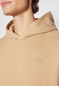 Sudadera beige con una textura suave, que presenta una capucha de canalé y un logo bordado en el pecho, ajustada para resaltar los detalles.