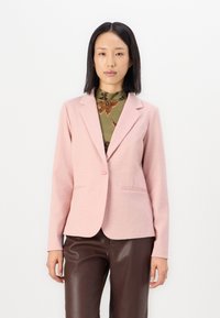 Světle růžový blazer vyrobený z měkké látky, s jedním knoflíkem, klopami a dvěma předními kapsami. Nositý přes vzorovaný zelený top.