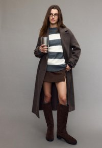 Žena s brýlemi, v dlouhém hnědém kabátě, pruhovaném svetru, hnědé mini sukni a vysokých hnědých botách, držící velký nerezový hrnek.