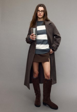 Ženska z očali, v dolgem rjavem plašču, črtastem puloverju, rjavi mini krilu in visokih rjavih škornjih, ki drži veliko steklenico iz nerjavečega jekla.