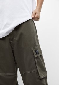 Mann trägt olivgrüne Cargo-Hosen mit einer Klappentasche und einem weißen Hemd, die Hand greift den Saum des Hemdes.