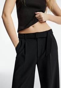 Black high-waisted trousers with a belted waist, pleats on the front, and side pockets. Made from smooth fabric, featuring tailored design.