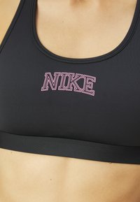Sujetador deportivo negro hecho de una tela suave que presenta una banda ancha y un logo "NIKE" en rosa en el centro. Escote redondeado con costuras mínimas.