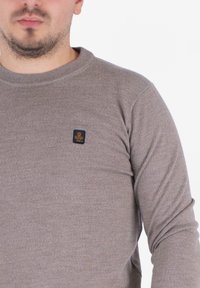 Uomo che indossa un maglione lavorato a maglia color marrone chiaro con un piccolo emblema nero e dorato sul petto sinistro, su uno sfondo bianco.