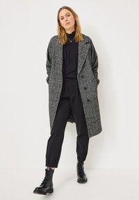 Manteau long texturé gris avec un design à double boutonnage, porté par-dessus un haut noir et un pantalon noir, associé à des bottes à lacets noires.