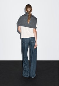 Grå stickad sjal runt axlarna över en vit t-shirt, i kombination med vida blå jeans. Bakgrunden är helt vit med mörkt golv.