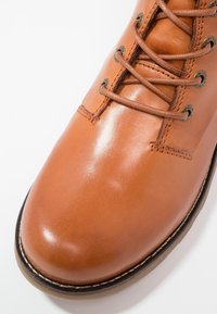 Gros plan d'une botte en cuir marron clair à lacets, avec un bout rond, des œillets métalliques et une couture détaillée le long de la semelle et de la tige.