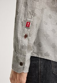 Close-up van een gestreepte grijze shirtmouw met bruine knopen en een rood etiket met een witte uitroepteken, gedragen met donkere jeans.