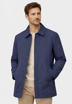 Uomo che indossa una giacca blu navy sopra un maglione beige e pantaloni color sabbia, in piedi con le mani nelle tasche della giacca, guarda di lato.