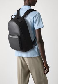 Schwarzer Rucksack aus glattem Material mit einer Fronttasche und verstellbaren Trägern, getragen über einem hellblauen Hemd und beigen Hosen.