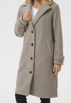 Kvinde iført en knælængde, tekstureret lyst gråt knaplukket frakke med brede reverser og forlommer, kombineret med sorte bukser og en hvid top.