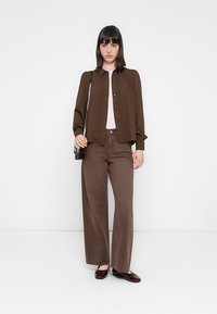 Camisa castanha de botão sobre uma blusa rosa claro, combinada com calças largas castanhas. O conjunto inclui uma mala preta e sapatos cor-de-vinho brilhantes.
