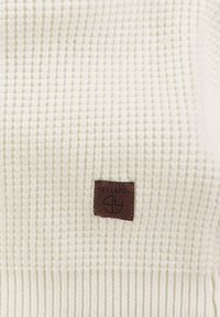 Witte gestructureerde brei-stof met een klein bruin leren "Key Largo" label in de hoek, voorzien van een ribgebreide rand aan de onderkant.