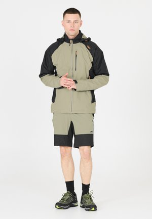 Uomo che indossa una giacca bicolore verde e nera con pantaloncini e calzini neri, in piedi con le mani incrociate, rivolto in avanti su sfondo bianco.