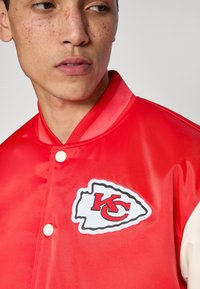 Κόκκινο σατέν μπουφάν με λευκή και μαύρη πινακίδα λογότυπου με το γράμμα "KC". Διαθέτει ριμπ γιακά και κλείσιμο με κουμπιά τύπου snap. Λείο υλικό.