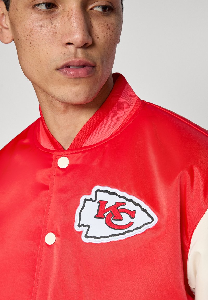 Rote Satinjacke mit weißem und schwarzem Logo-Patch mit der Aufschrift "KC". Verfügt über einen gerippten Kragen und einen Druckknopfverschluss. Glatte Textur.