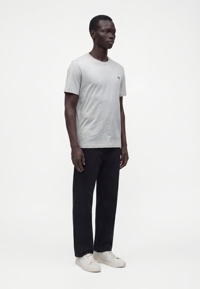 Homme grand debout face à l'avant portant un t-shirt gris clair à manches courtes, un pantalon noir et des baskets blanches sur un fond blanc uni.