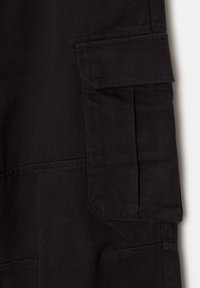 Gros plan sur un tissu noir montrant une poche cargo avec un rabat et des coutures visibles sur un pantalon ou un short.