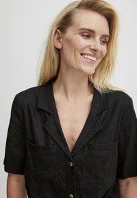 Juodas trumpomis rankovėmis marškinėliai su išpjauta apykakle, dviem priekinėmis kišenėmis ir sagų uždarymu. Audinys atrodo tekstūruotas ir lengvas.