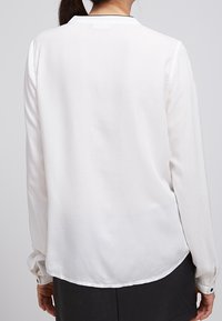 Blouse blanche à manches longues en tissu léger, avec un ourlet arrondi et des poignets à boutons. Design simple, texture lisse.