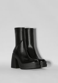 Botines de cuero negro con un acabado brillante, tacones de bloque altos y angulares, y suelas gruesas. Diseño minimalista con acentos de costura.