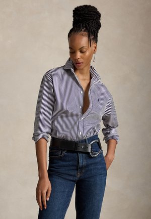 Femme avec un chignon tressé portant une chemise boutonnée à rayures bleu et blanc, un jean foncé et une ceinture noire, debout avec une main dans la poche.