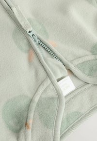 Fleece'owa kurtka zapinana na zamek błyskawiczny w jasnym zielonym kolorze, z pastelowymi kropkami. Posiada metalowy zamek i miękką teksturę materiału. Metka widoczna w środku.