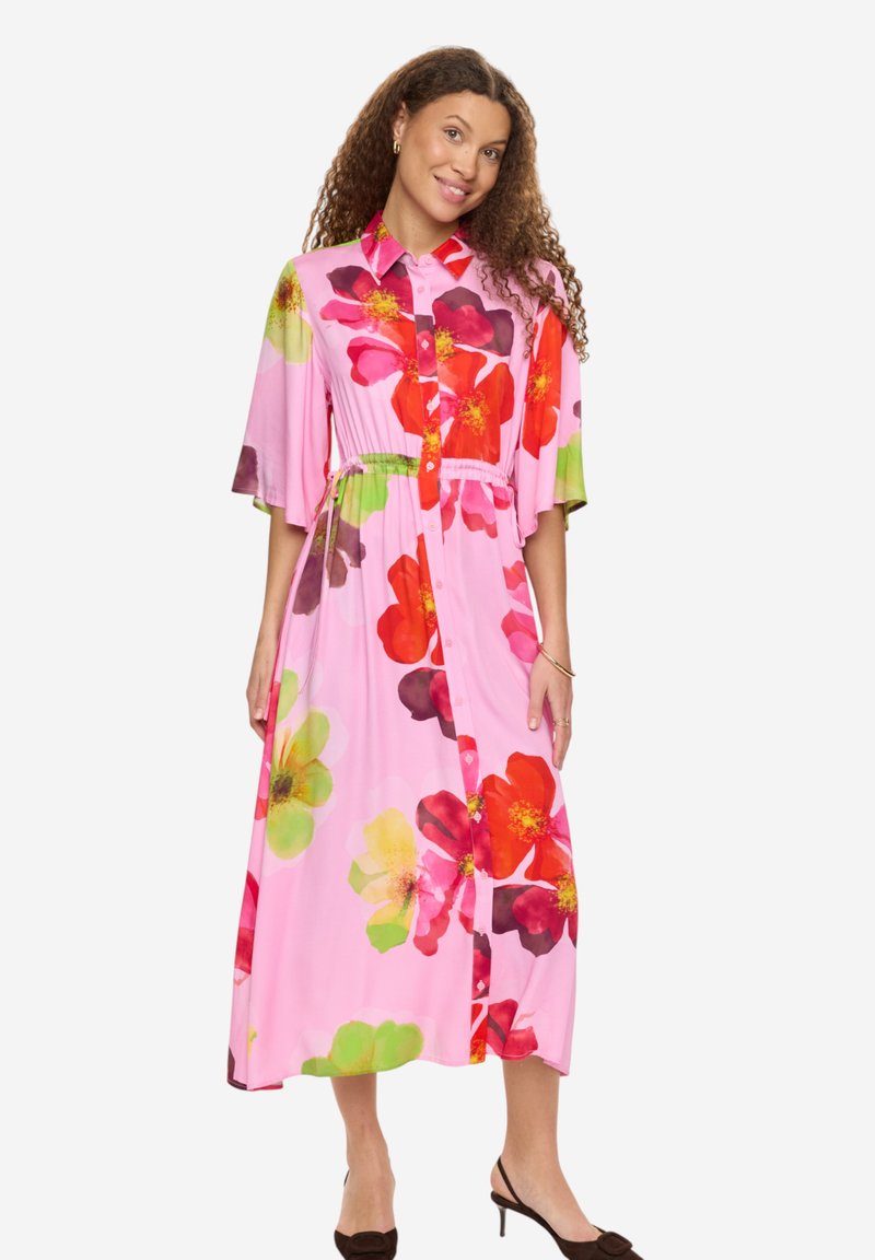 Femme portant une robe midi rose avec de grands imprimés floraux rouges, verts et jaunes, boutonnée à l'avant, manches trois-quarts, debout avec un sourire.