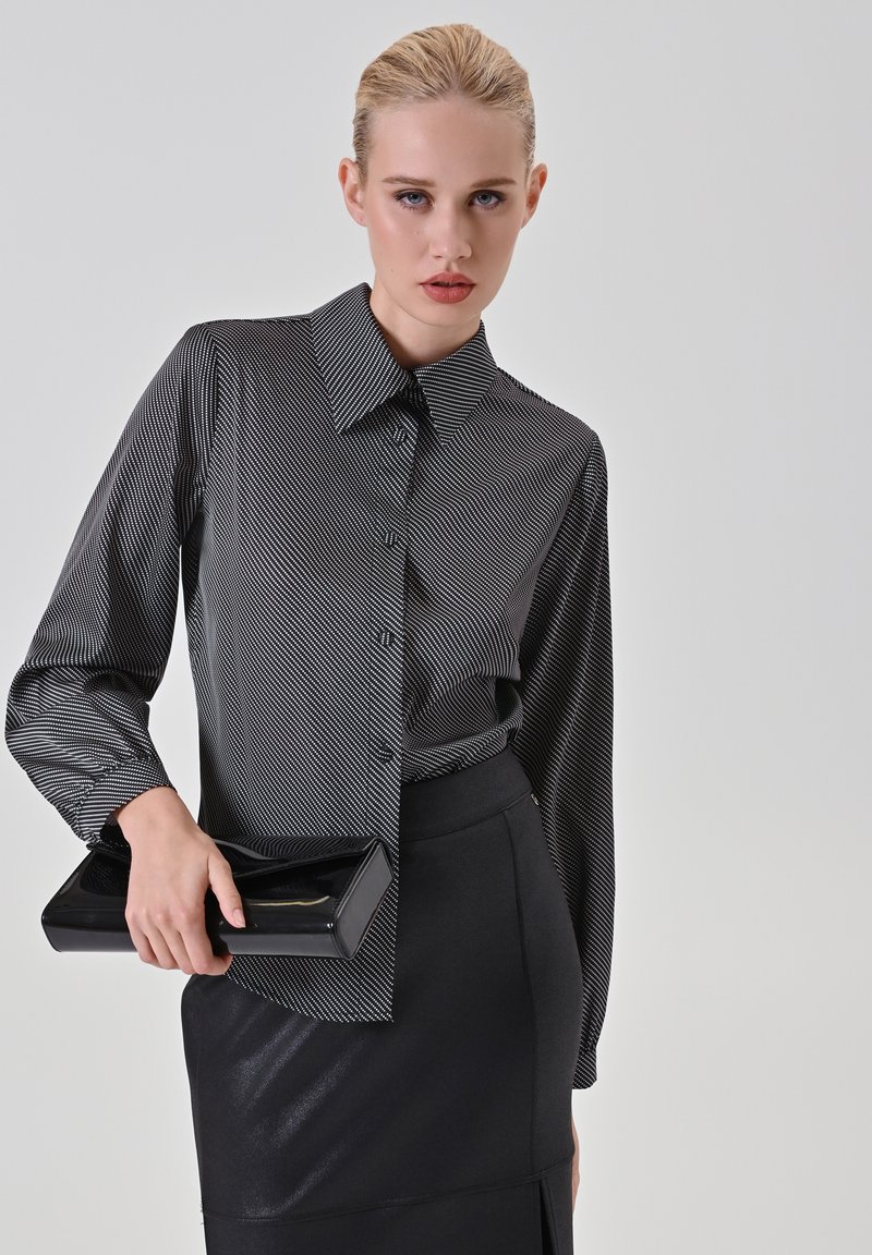 Blusa a righe bianche e nere con colletto alto, maniche lunghe e bottoni frontali, abbinata a una gonna a tubino nera aderente e una pochette lucida nera.