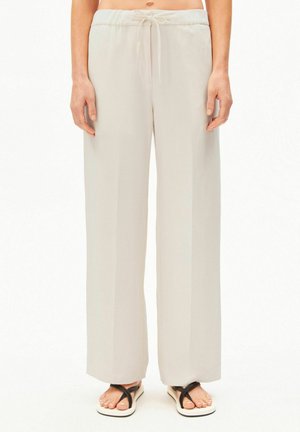 Frau trägt lockere, hellbeige Hosen mit Kordelzug und weiße Sandalen mit schwarzen Riemen, steht vor einem schlichten weißen Hintergrund.