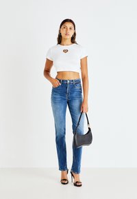 Λευκό κοντό μπλουζάκι με κομμένο σχέδιο σε μορφή καρδιάς, μπλε εφαρμοστά τζιν, μαύρες ανοιχτές γόβες και μια υφασμάτινη μαύρη τσάντα. Ουδέτερο φόντο.