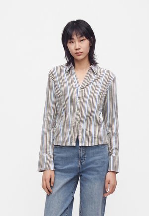 Jeune femme portant une chemise boutonnée à manches longues avec des rayures verticales et un jean bleu sur un fond clair uni.