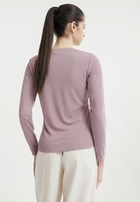 Langärmliges Oberteil aus weichem Mauve-Stoff mit gerippter Textur, ausgestattet mit einem Rundhalsausschnitt und taillierter Passform, kombiniert mit cremefarbenen Hosen.