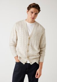 Junger Mann, der eine cremefarbene, strukturierte Strickjacke über einem weißen Hemd und einer dunklen Jeans trägt, steht vor einem neutralen Hintergrund.