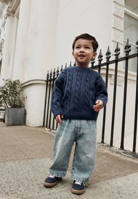 Gebreide marineblauwe trui met kabelpatroon, gecombineerd met losse lichtblauwe denim jeans en marineblauwe sneakers met witte veters, staand op een trottoir.